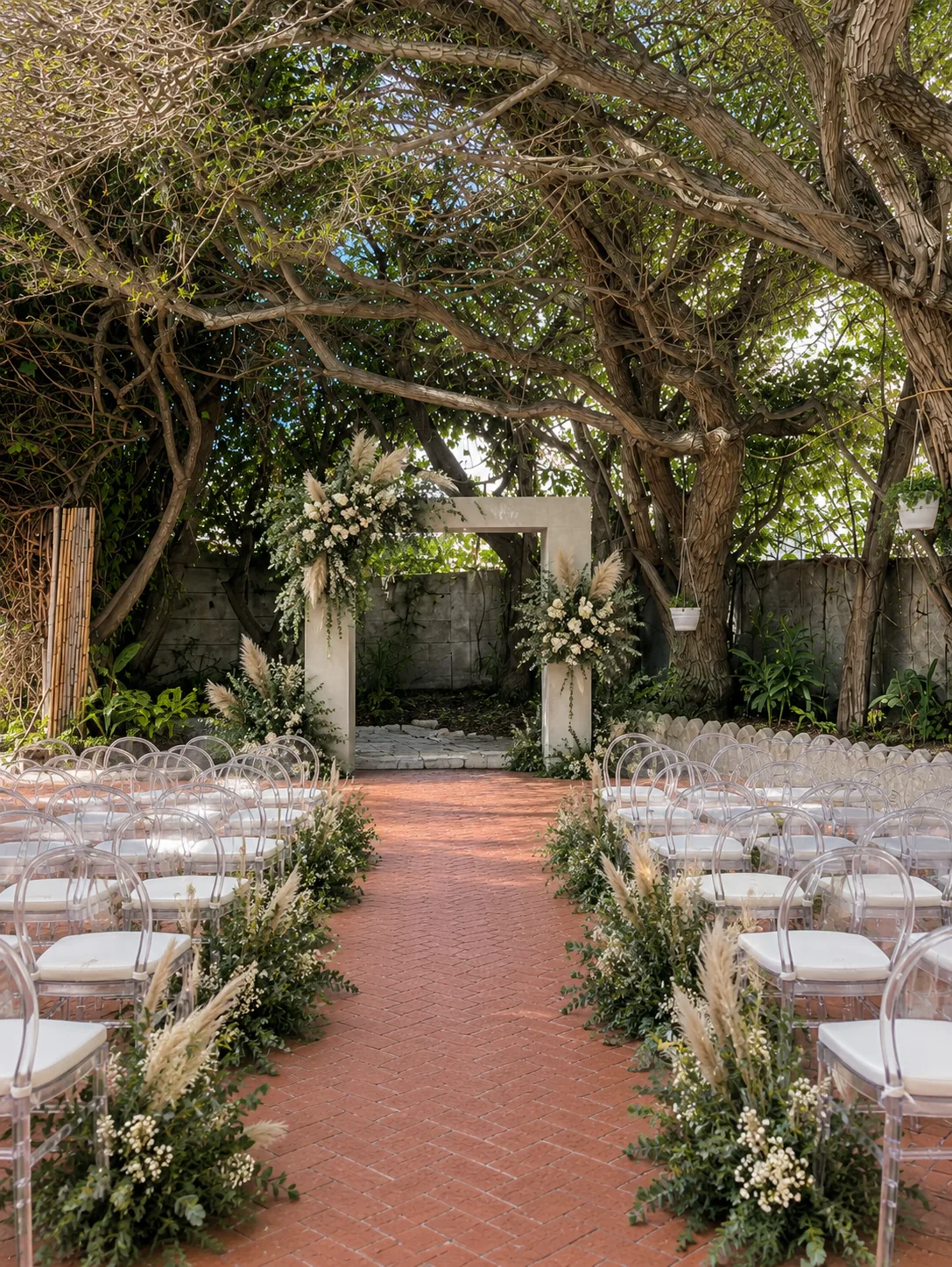 beach-collection-garden-wedding-ceremony-arch Outdoor wedding ceremony aisle with clear chairs, greenery and a white floral arch beneath mature trees.